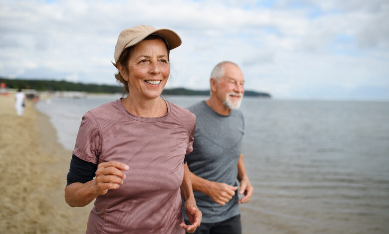 Active senior couple runners jogging outdoors on sandy beach by sea in early morning.